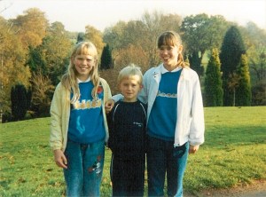 Three children stand together, smiling, in front of trees