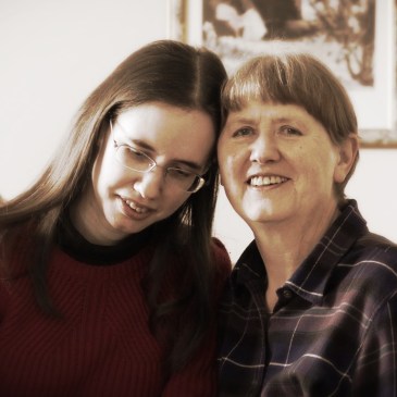 Two women sit with their heads resting supportively together. One looks slightly downwards. The other smiles at the camera.
