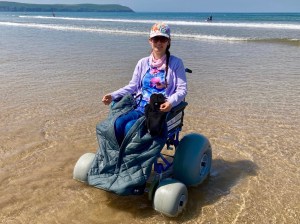 Naomi sits at the edge of the sea in a beach wheelchair (which has special large wheels for the sand). She wears sunglasses and a multi-coloured cap, and is smiling at the camera. She is wearing a blue and pink top with a flower pattern, and a pale purple cardigan. Her jeans are partially covered by a wheelchair leg cover. It is a bright, sunny day.