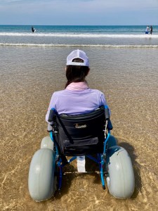 Naomi sits in a wheelchair at the edge of the sea. The camera is behind her. The sea and sky are blue.