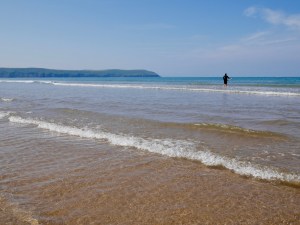 The sea as seen from the shore, looking out towards the horizon and headland. The sea is clear and sparkling in the foreground, and blue further out. A surfer stands in the waves.