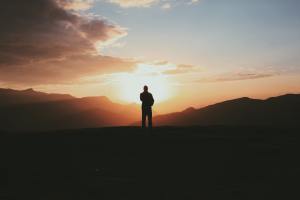 A silhouetted figure stands looking out across a mountain range. The sun is rising, and the peaks of the mountains are also in shadow.