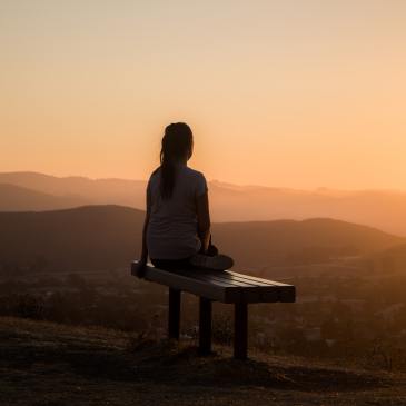 A silhouetted figure sits on a bench, looking out over hills.  The sun is setting over the scene.
