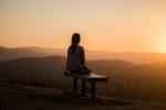 A silhouetted figure sits on a bench, looking out over hills. The sun is setting over the scene.