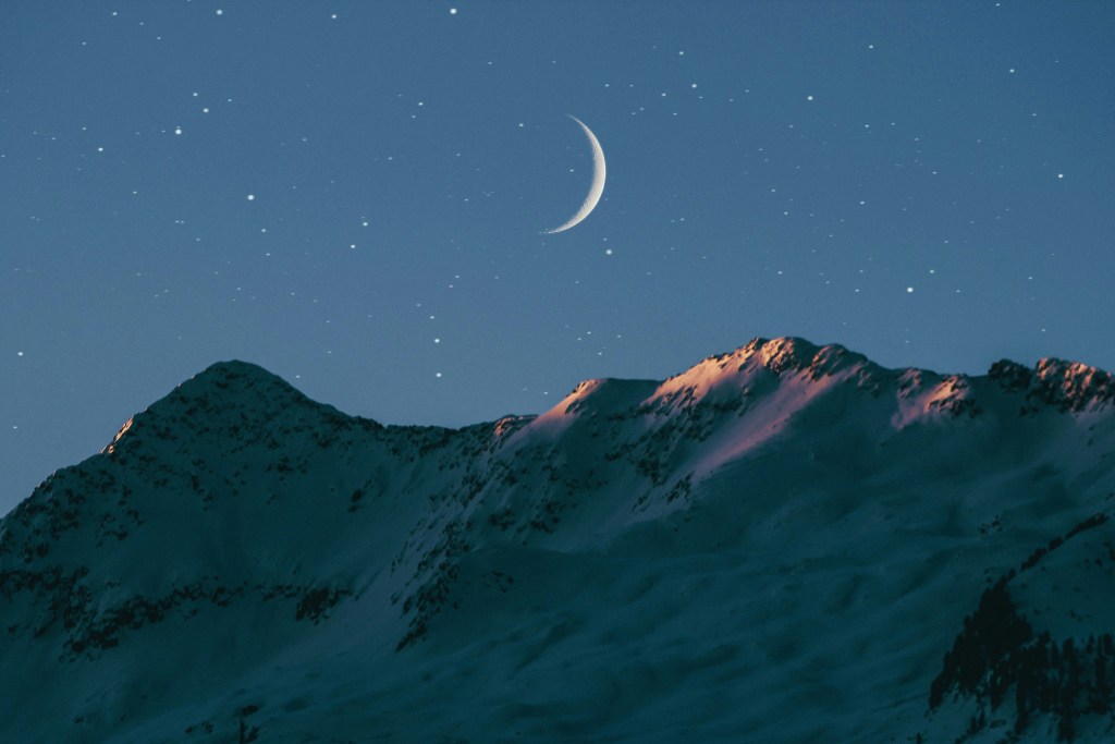 A crescent moon in a starlit sky, over a snowy mountain