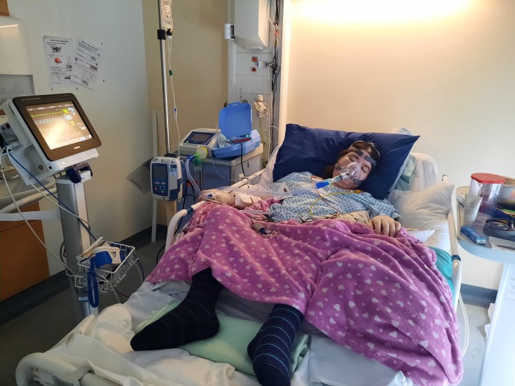 Kara lies seriously ill in a hospital bed. She has an oxygen mask and a drip.