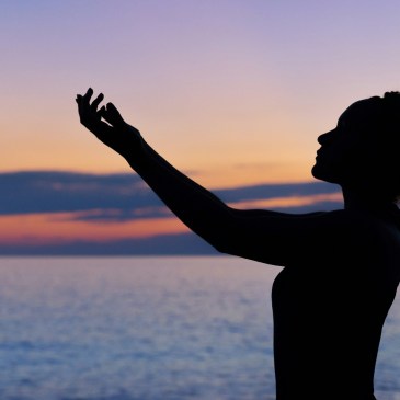 A silhouetted image of a woman, standing in front of the sea and a sunset sky. Her arms are outstretched in front of her, as though in thanks or pleading.