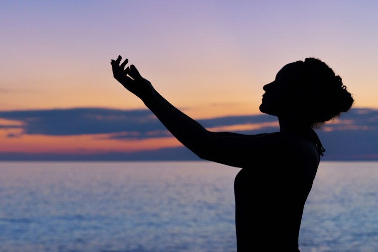 A silhouetted image of a woman, standing in front of the sea and a sunset sky. Her arms are outstretched in front of her, as though in thanks or pleading.